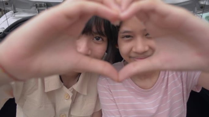 两个亚洲开朗可爱的女孩一起做手势心形，坐在掀背车的后侧看着相机。年轻的生活方式。