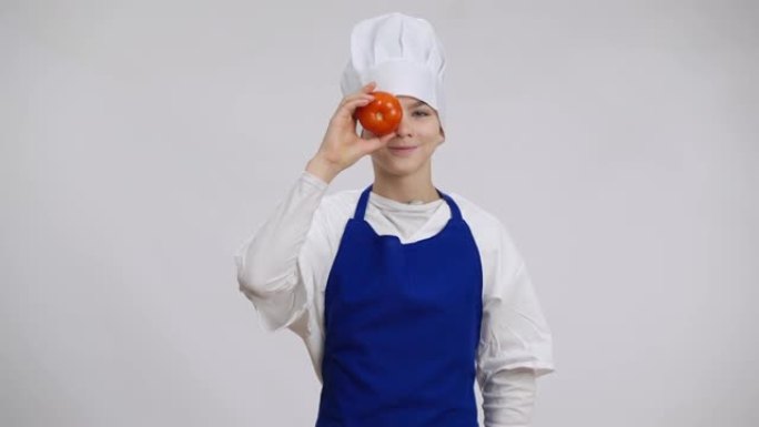 穿着厨师制服的积极英俊的高加索男孩与西红柿合眼。欢快可爱的小厨师在白色背景下看着相机享受食物的肖像