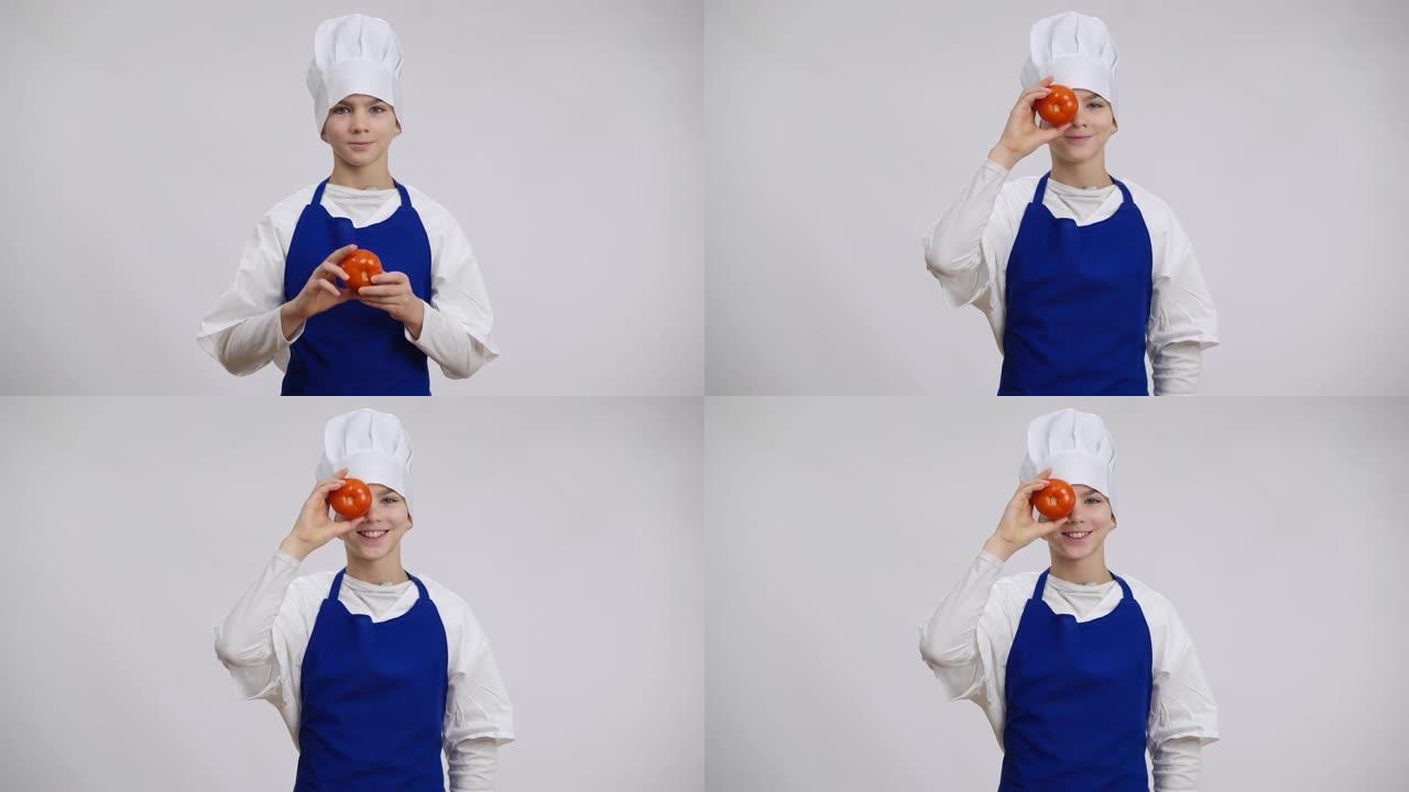 穿着厨师制服的积极英俊的高加索男孩与西红柿合眼。欢快可爱的小厨师在白色背景下看着相机享受食物的肖像