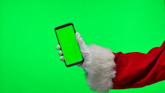 圣诞老人戴着白手套的手将智能手机放在垂直位置，并带有绿色屏幕色度键。孤立在工作室的绿色背景上。模拟显