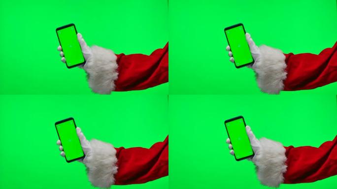 圣诞老人戴着白手套的手将智能手机放在垂直位置，并带有绿色屏幕色度键。孤立在工作室的绿色背景上。模拟显