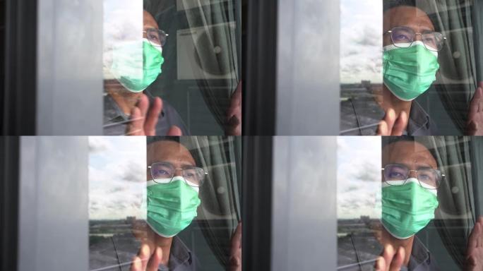 一名亚洲男子戴着防护口罩透过阳台窗户向外看
