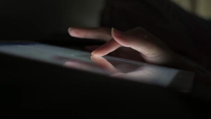 一个女人的手指在平板电脑的屏幕上滑动，可以在不打开灯的情况下播放社交媒体，在睡觉前躺在床上。