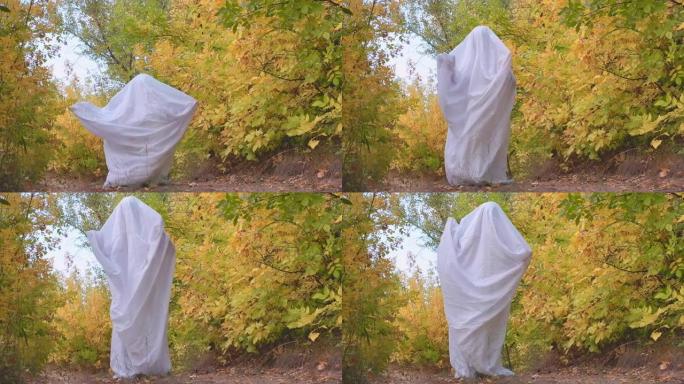 秋天森林里一个穿着幽灵服装的男人在下午庆祝万圣节。鬼魂鬼混，拍拍手，穿着白纸玩得开心