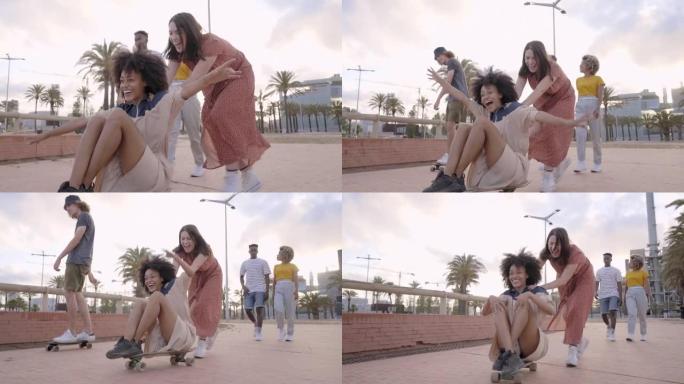 一群年轻人在城市里玩滑板。坐在滑板上微笑的黑人女孩被白人女孩推拉。女孩。