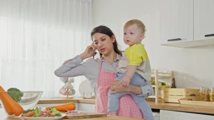 高加索人忙碌的母亲和男婴蹒跚学步的孩子在厨房做家务。美丽的单身妈妈用电话上班和做饭为家里的小孩准备早