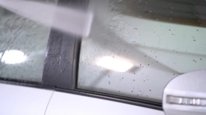水雾清洗一辆白色汽车。关闭车库部分侧门玻璃上的喷水清洗。在车辆上使用喷水喷雾的工人。
