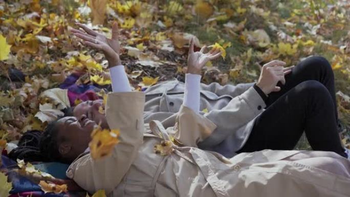 混合赛跑的夫妇在秋天躺在公园的毯子上，向空中扔树叶