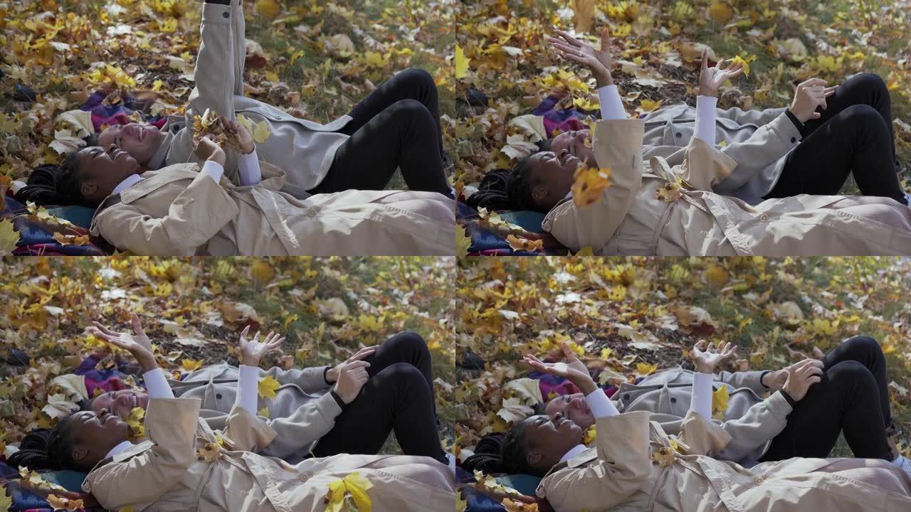 混合赛跑的夫妇在秋天躺在公园的毯子上，向空中扔树叶