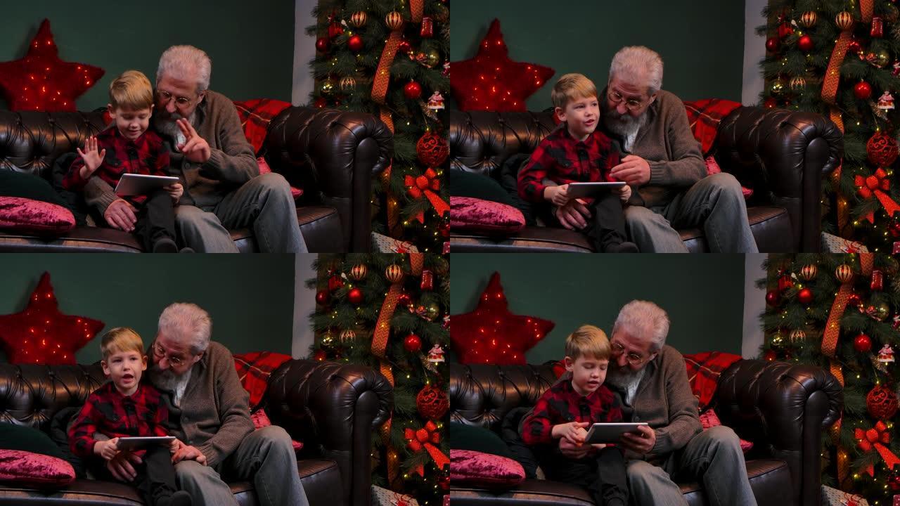 优雅的老人和小男孩在平板电脑上谈论视频交流。祖父和孙子坐在靠近发光圣诞树的装饰房间的沙发上。特写。慢