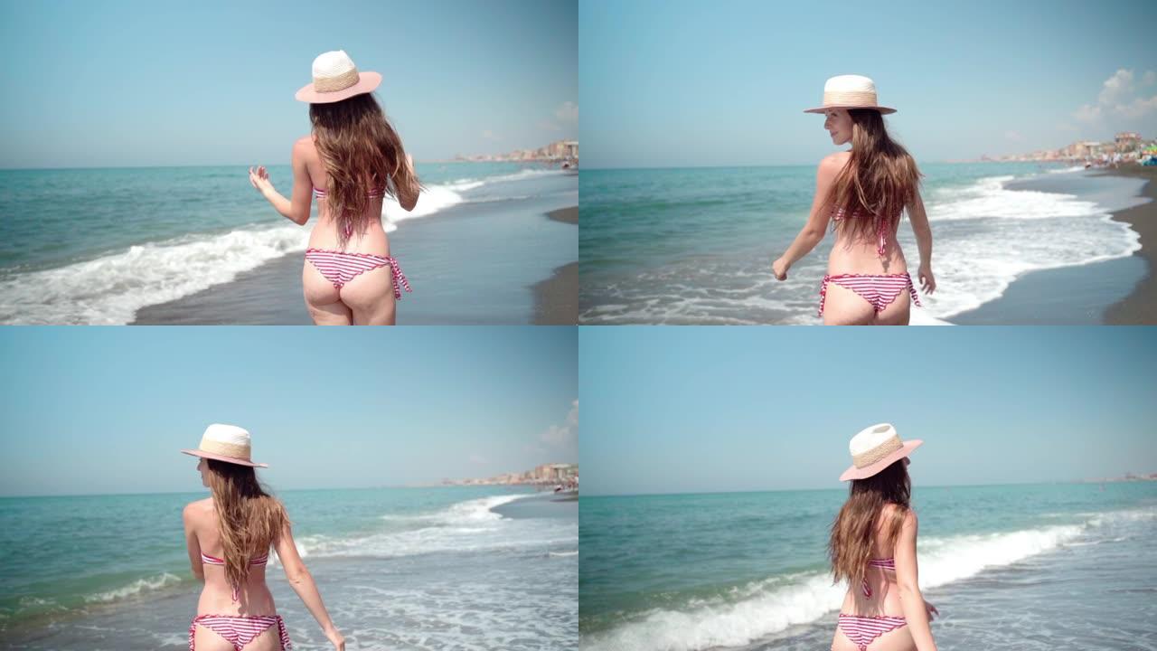 迷人的白人黑发女人穿着夏天的帽子和性感的比基尼泳装在海边慢动作行走，转身，玩弄头发和微笑。苗条的女游
