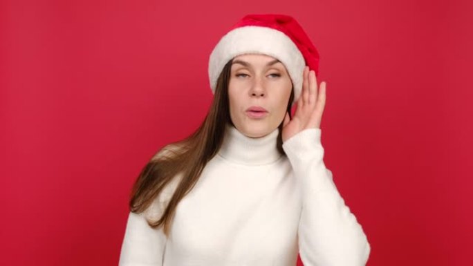 穿着白色舒适毛衣和圣诞老人圣诞帽的获胜者年轻女子试图听到你偷听的声音，手靠近耳朵，孤立在红色工作室背