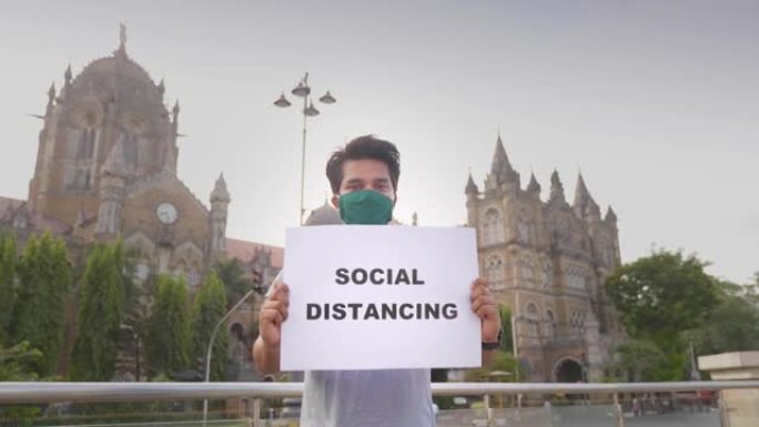 一个戴着口罩的年轻人站着，举着标语 “社交距离”