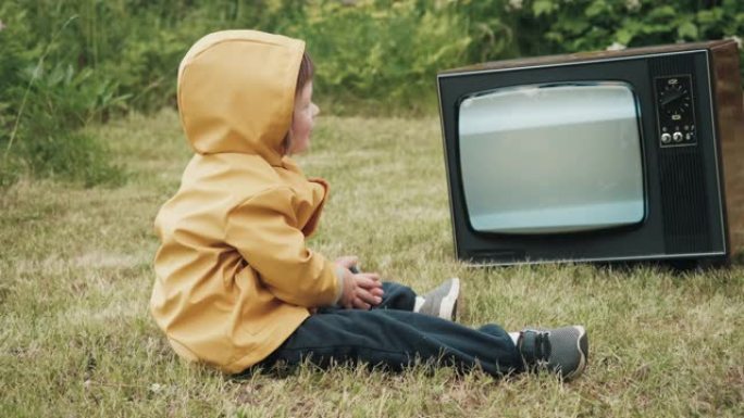 孩子穿着带兜帽的黄色雨衣，正在看复古电视，微笑着