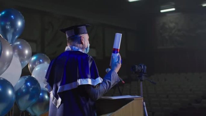 在线毕业，大学讲师穿着学术服装和戴着医用口罩的帽子在会议室使用摄像机颁发文凭