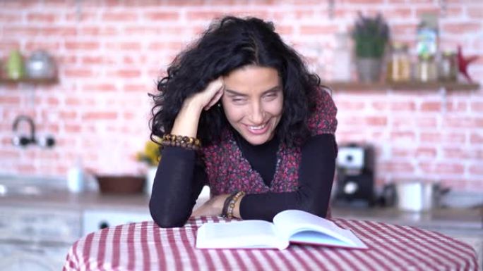 4k视频笑着卷曲的黑发女人坐在厨房的桌子上，在家看书