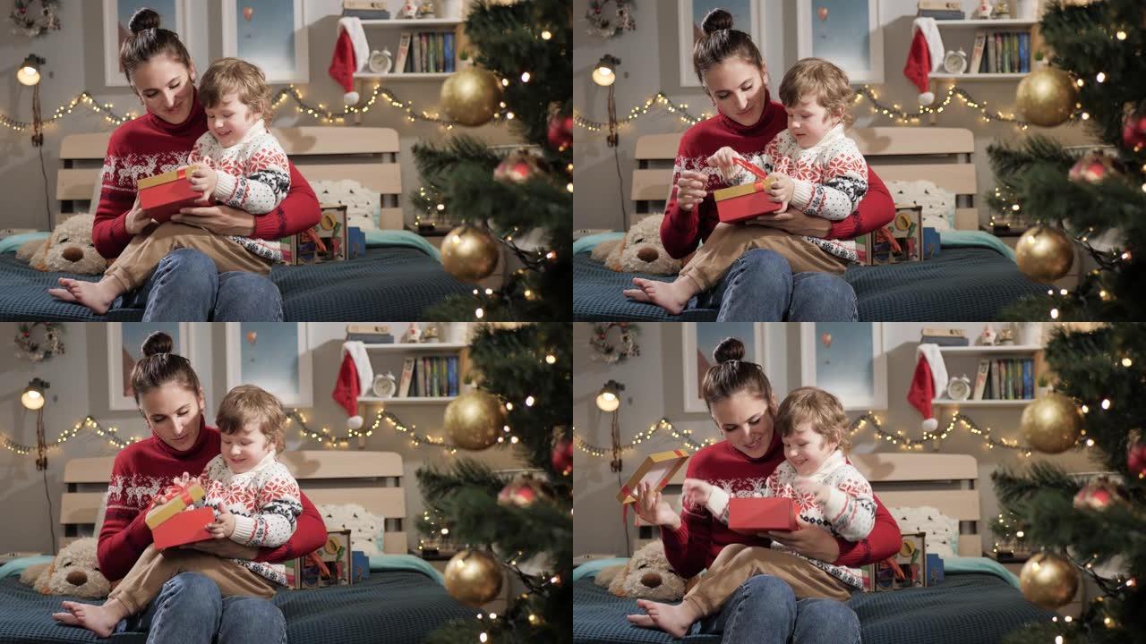 孩子打开圣诞礼物。家里2-3岁的母亲和孩子坐在床上，婴儿打开圣诞礼物，欢欣鼓舞，鼓掌和欢笑。慢动作