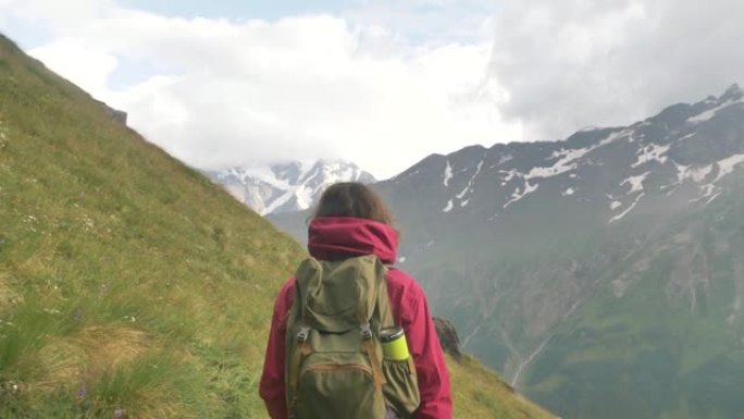 一位女游客背着背包，在白雪皑皑的山峰的背景下，沿着山路上的绿色斜坡行走