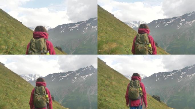一位女游客背着背包，在白雪皑皑的山峰的背景下，沿着山路上的绿色斜坡行走