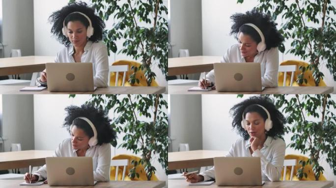 使用笔记本电脑和耳机在记事本上做笔记的非裔美国妇女在视频电话会议上讲话的肖像。布鲁内特坐在家庭办公室