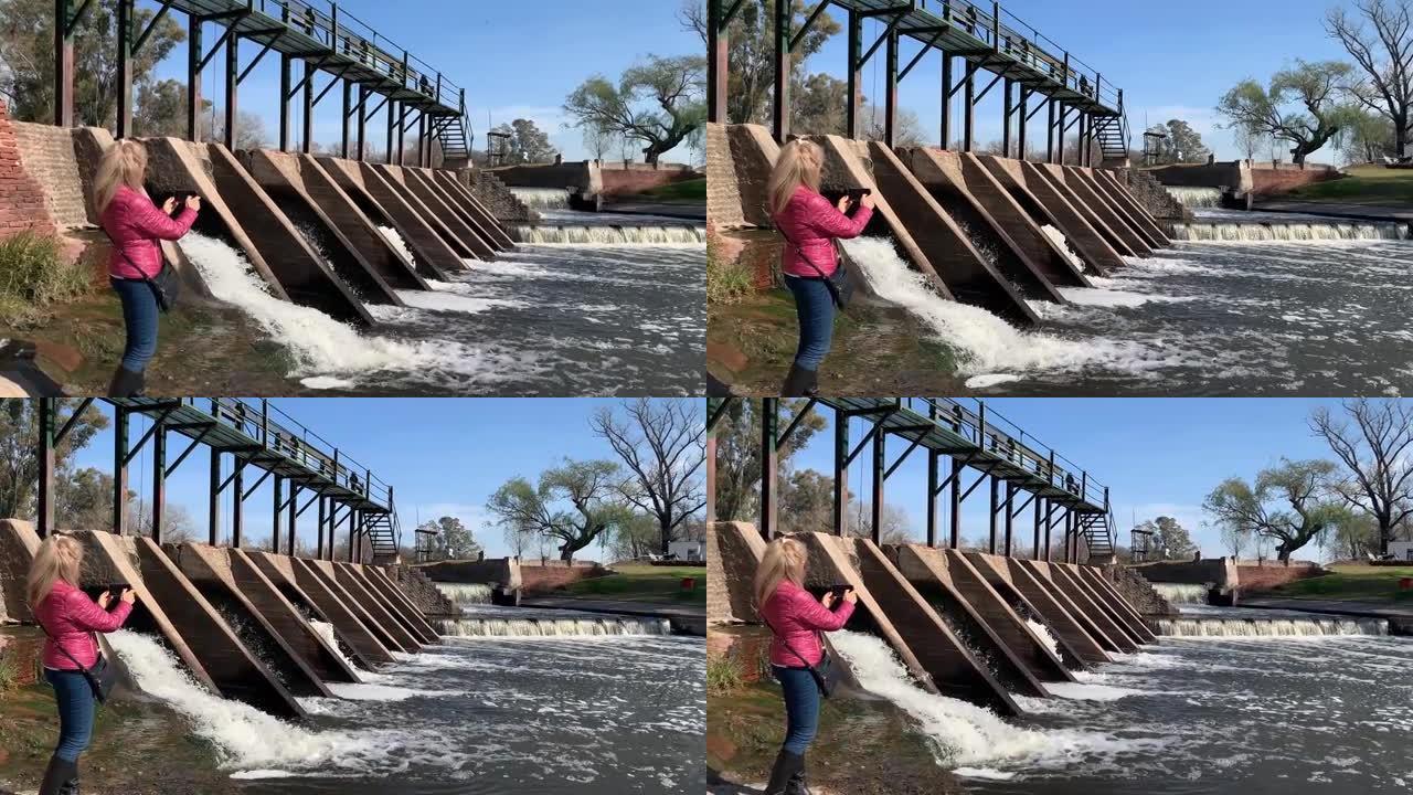 金发女子在大坝大门的水流出处用手机拍照。圣安东尼奥·德·阿雷科。旅行概念