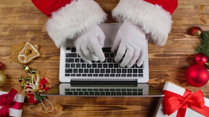 顶视图圣诞老人戴着白手套在木制新年装饰桌子上键盘打字。圣诞老人与笔记本电脑一起工作，浏览邮件并回答给