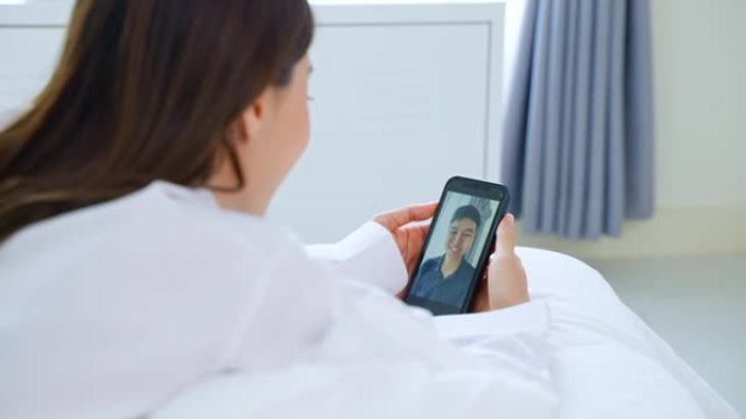 亚洲美女在家床上和男朋友视频通话。穿着睡衣的迷人女孩早上在卧室醒来后，喜欢在手机上的虚拟遥控器上与年