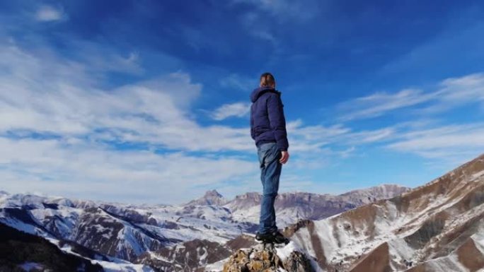鸟瞰年轻的高加索大胡子的时尚男子站在一块大石头上的风景如画的雪山岩石。晴天，蓝天有云