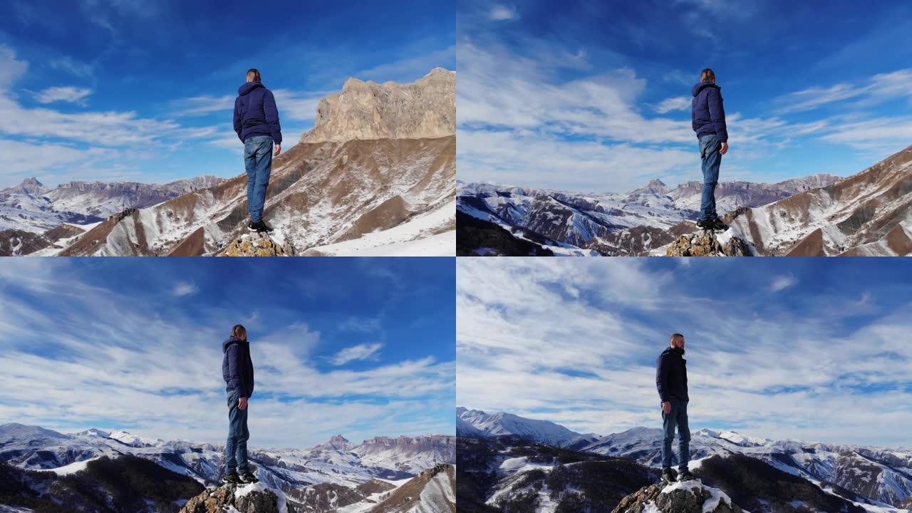 鸟瞰年轻的高加索大胡子的时尚男子站在一块大石头上的风景如画的雪山岩石。晴天，蓝天有云