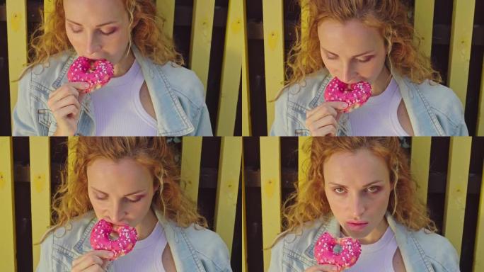美丽的年轻女子开胃咬掉粉红色的甜甜圈。红色卷发女孩，有着不同寻常的黄色眼睛，吃甜食。穿着牛仔裤夹克的