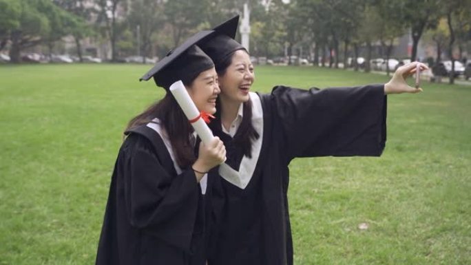慢动作快乐的一群成熟的学生在毕业日摆姿势自拍。两个穿着学术服装和帽子的亚洲最好的女朋友自拍。微笑迷人
