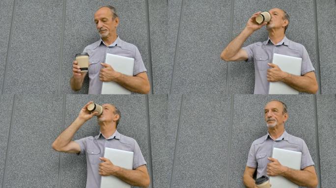 年长同事的男性肖像，带着银色的笔记本电脑和一杯咖啡，早上在深灰色的墙壁附近等待会议，远程工作，喝咖啡