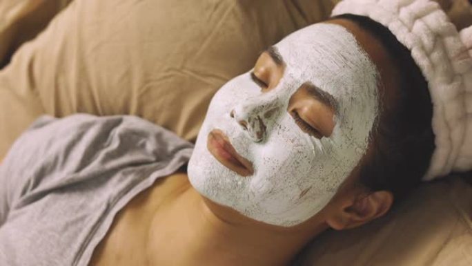 用泥遮住脸的中等年龄亚洲女性的面部聚焦显示她闭着眼睛躺在床上，放松温暖的光线，在水疗中心听音乐，享受