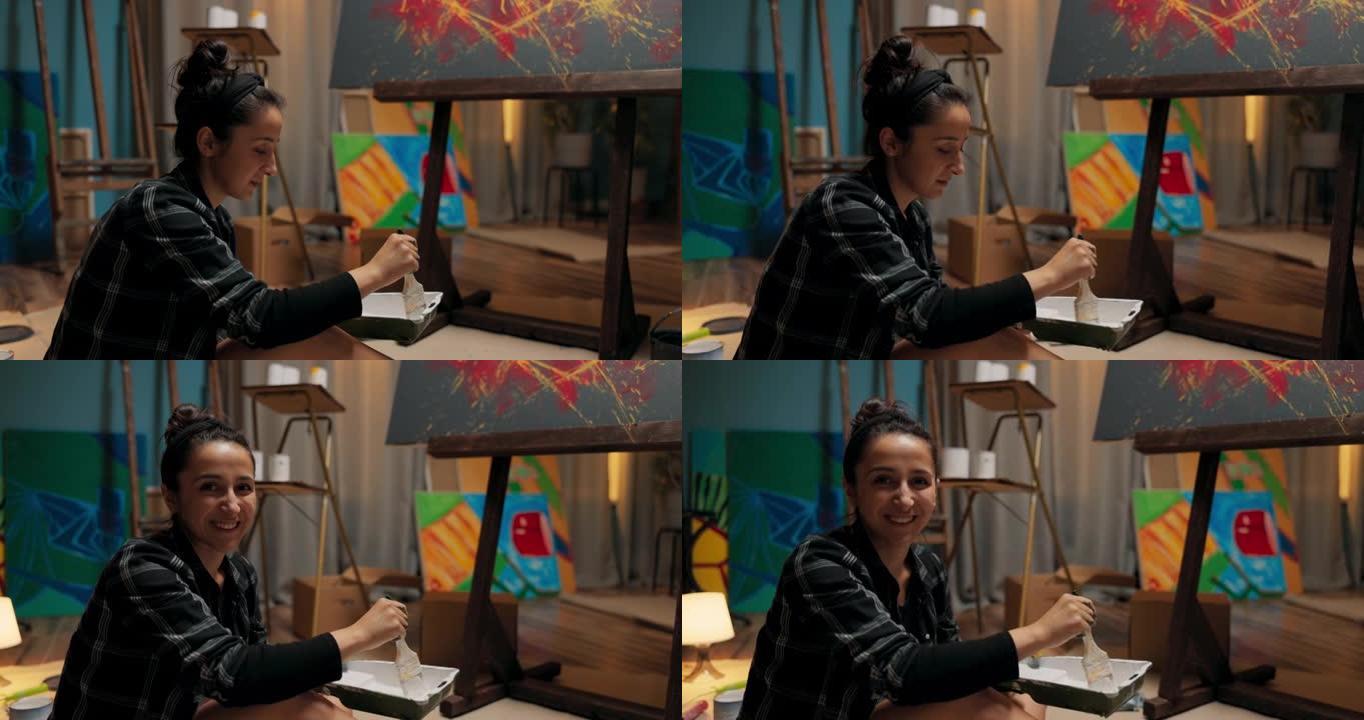 一位年轻的艺术家坐在用纸板固定的地板上，在油漆和绘画中，拿着刷子。尽管她的手很脏，但这个女人仍然对自