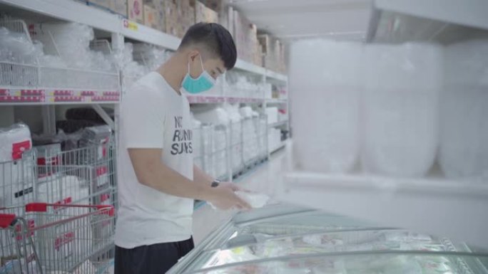 一名年轻的亚洲男子打开玻璃滑门，在超市的冰柜里拿走一包冷冻食品，买一送一促销，省钱，食品行业的技术，