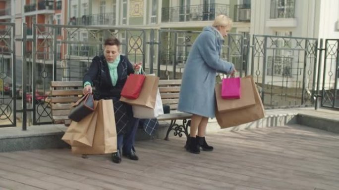 老年女性买家带着许多纸袋在户外搓揉酸痛的脚
