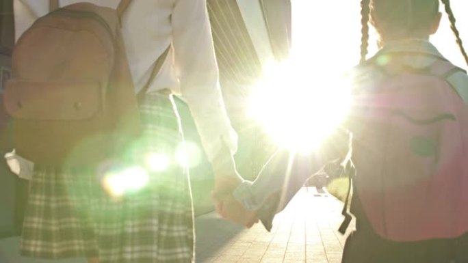 两个不同年龄的女学生背着背包手牵着手从学校回来。姐姐帮助年轻人。后视图