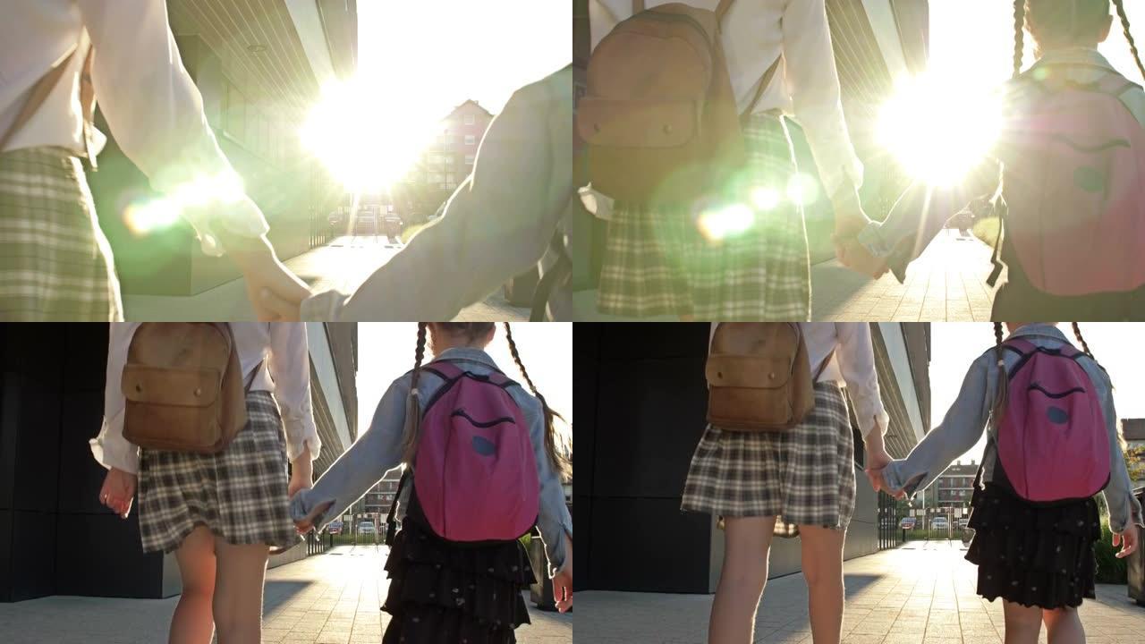 两个不同年龄的女学生背着背包手牵着手从学校回来。姐姐帮助年轻人。后视图