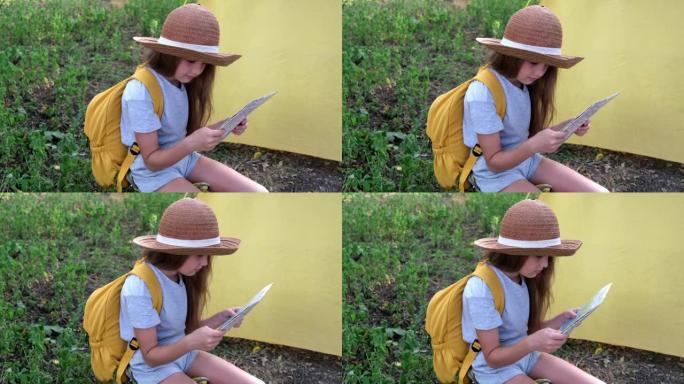 戴着帽子，背着黄色背包的小女孩坐在帐篷附近的夏令营里。孩子在地图上移动手指。儿童地图阅读技能的概念。