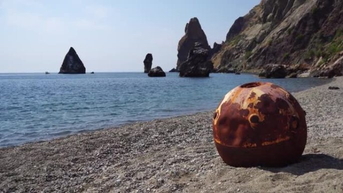 陈旧生锈的圆形漂浮在海滩上的水雷与岩石海岸和海洋背景。污染、保护自然、战争与和平的理念