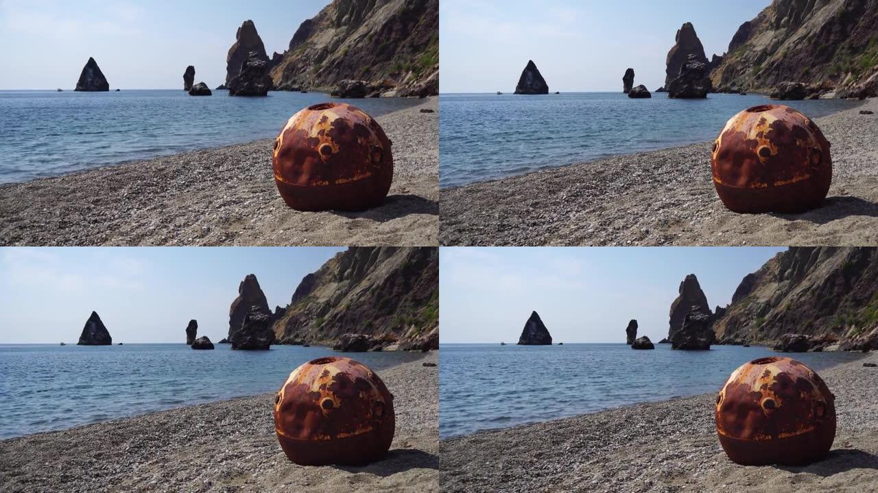 陈旧生锈的圆形漂浮在海滩上的水雷与岩石海岸和海洋背景。污染、保护自然、战争与和平的理念