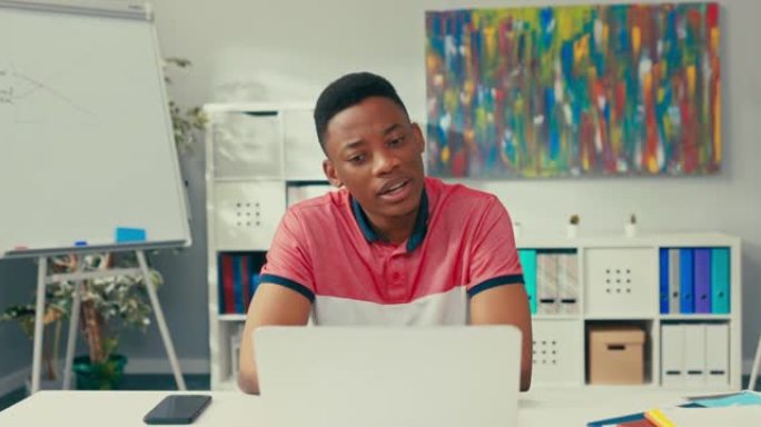 一个非洲裔美国美女的小男孩坐在办公室里的桌子上，拿着一台笔记本电脑，面带微笑，挥手致意，通过互联网进
