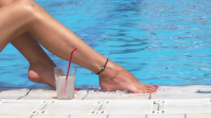 性感的女腿在公共泳池上，用塑料吸管端着一杯鸡尾酒或柠檬汁