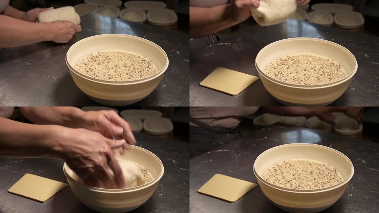 将一块生面团浸入装有种子和谷物的碗中的女性手的特写镜头。烤面包前准备好面团。工艺面包店