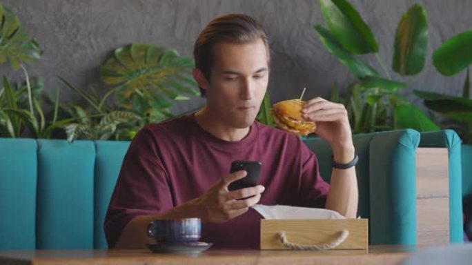 男人在小咖啡馆喝咖啡休息时吃汉堡和滑动智能手机