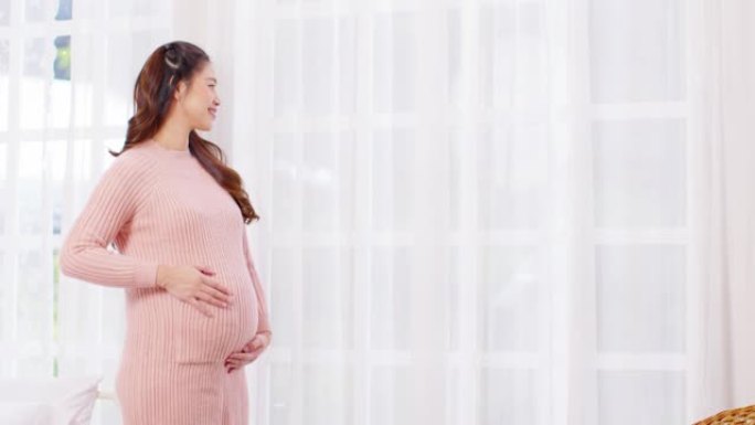 快乐的孕妇站在窗前，在舒适的家中用爱抚摸她的大肚子，年轻女子的怀孕享受未来的生活，用爱，母性和怀孕的