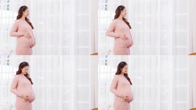 快乐的孕妇站在窗前，在舒适的家中用爱抚摸她的大肚子，年轻女子的怀孕享受未来的生活，用爱，母性和怀孕的