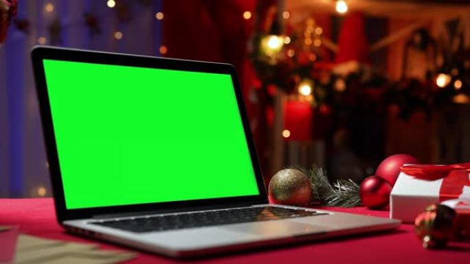 带有绿屏色度键的打开笔记本电脑站在礼物和玩具旁边的红色桌子上。家庭房间，圣诞装饰和灯光在模糊的背景。