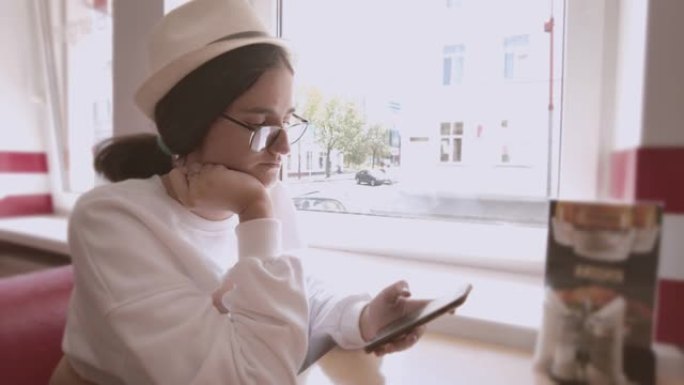 戴着帽子和眼镜的女孩坐在咖啡馆里，用手机，等待订单，跟踪摄像头