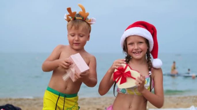 沙滩上戴着圣诞老人帽子和鹿角的孩子。小男孩和女孩在跳舞，微笑着看着镜头。圣诞快乐。寒假。大海在背景中